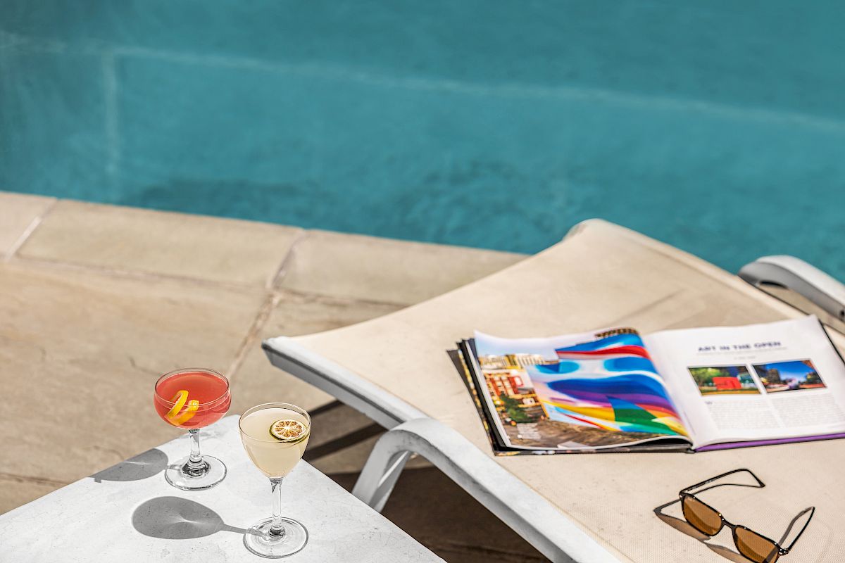 Two cocktails on a table by a pool, with a magazine and sunglasses on a lounge chair.